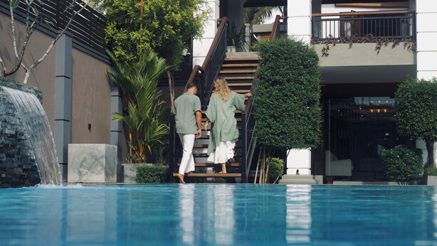 Una piscina con caída de agua, rodeada de vegetación y un edificio moderno con una escalera.