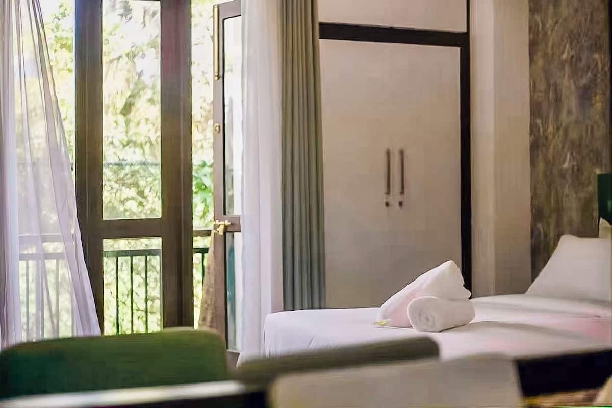 A hotel room with a bed, a green chair, a wardrobe, and a view of greenery outside.
