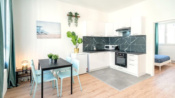 A modern kitchen with white cabinets, a black countertop, and a marble backsplash. There is a dining area with a table and chairs, and a bed in the adjacent room.
