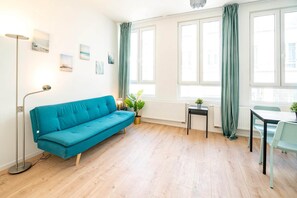 A modern living room with a teal sofa, a floor lamp, a dining table with chairs, and large windows with curtains.