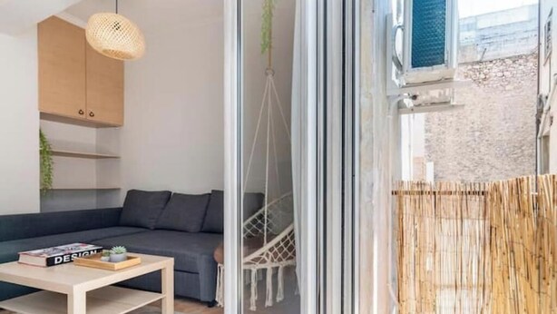 A modern living room with a sofa, a coffee table, a hanging light fixture, and a view of a balcony with a wicker fence. - STM Monastiraki Acropolis Apt (Athina)