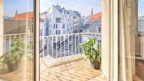 Un balcon orné d’un pot de fleurs, une vue sur une rue de la ville avec ses bâtiments, et un ciel dégagé.