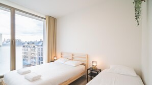 Una camera da letto con un'ampia finestra, un letto con lenzuola bianche, un comodino con una lampada e vista sugli edifici.