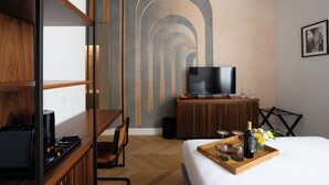 A modern hotel room with a wooden desk, a flat-screen TV, a bed with a tray of wine glasses and flowers, and a unique wall design. - Atelier Alinari Rooms (Florence)