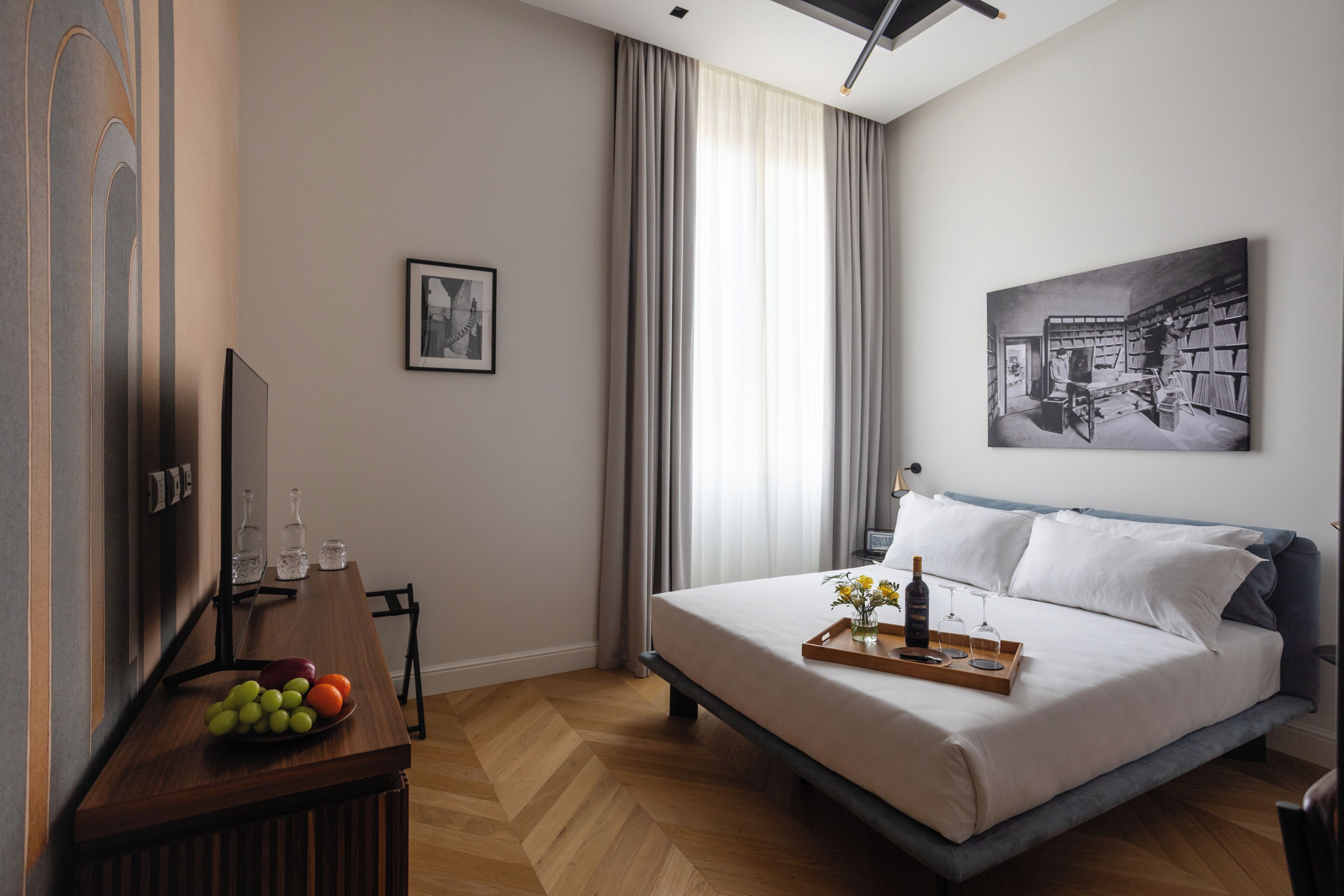 A hotel room with a bed, a dining table with fruit, a TV, and a painting on the wall.