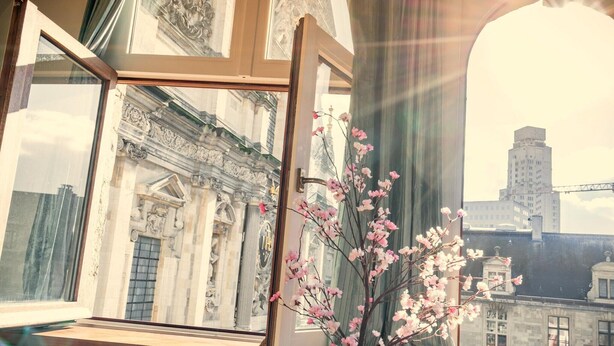 Ein offenes Fenster mit Blick auf eine Gebäudefassade und einer Blumenvase.