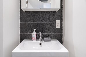 Un baño con un lavabo blanco, salpicadero de azulejos oscuros, un espejo sobre el lavabo y una botella de jabón para manos.