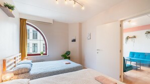 Une chambre avec deux lits, une fenêtre avec des rideaux, une plante et une vue sur un canapé bleu situé dans la pièce adjacente.