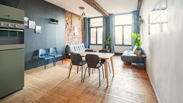 A modern kitchen with a built-in oven, a dining area with a wooden table and chairs, and a seating area with a sofa. - Spacious riverside apartment for 9 (Antwerp)