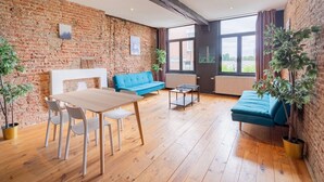 Una habitación con pared de ladrillo, suelo de madera, un sofá azul, una mesa de madera con sillas y una ventana con cortinas.