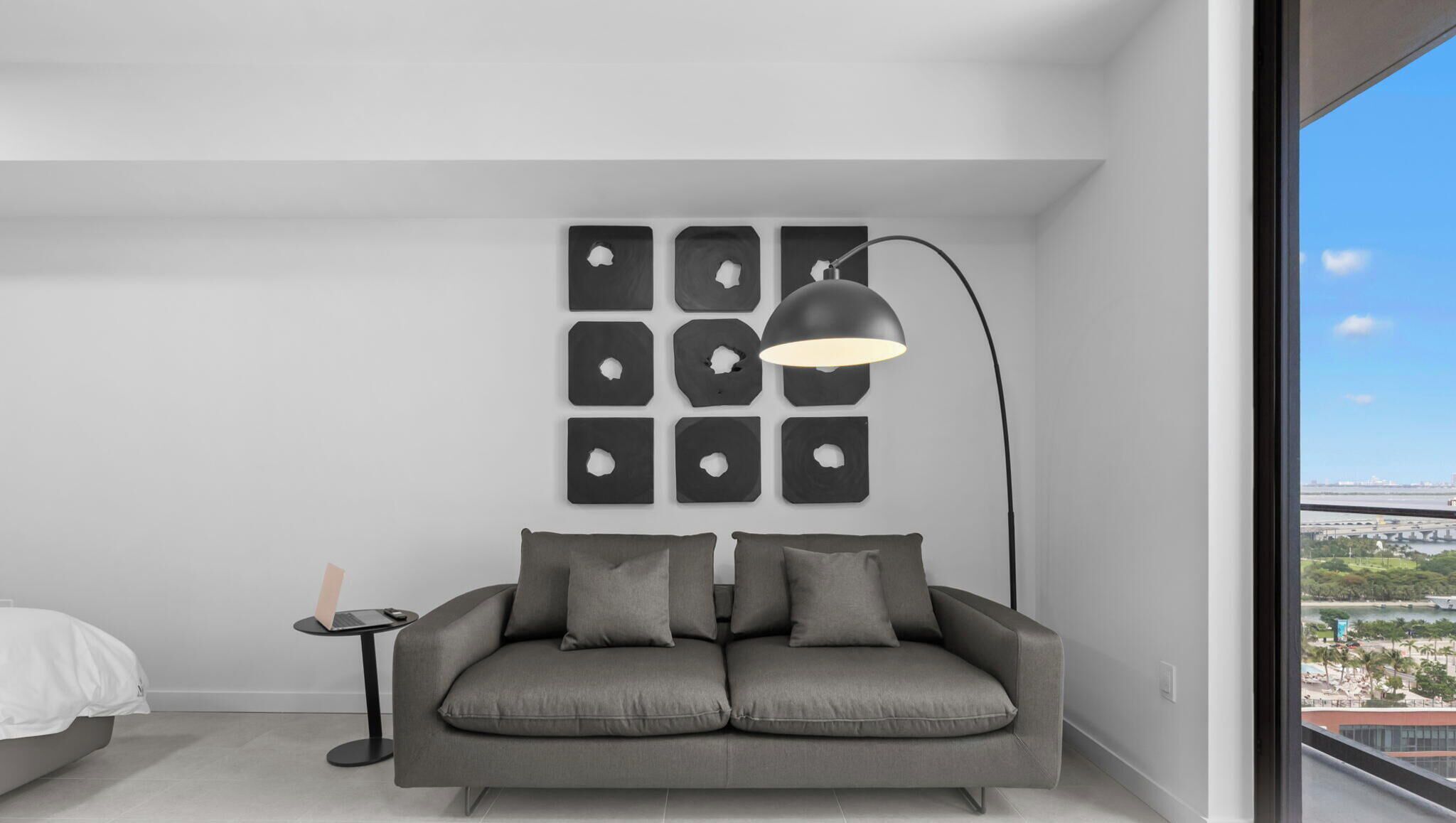 A modern room with a grey sofa, a black floor lamp, and a city view through a large window.