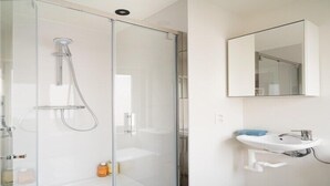 Uma casa de banho moderna com cabine de duche em vidro, uma sanita branca com espelho em cima e uma pequena prateleira com produtos de higiene pessoal.