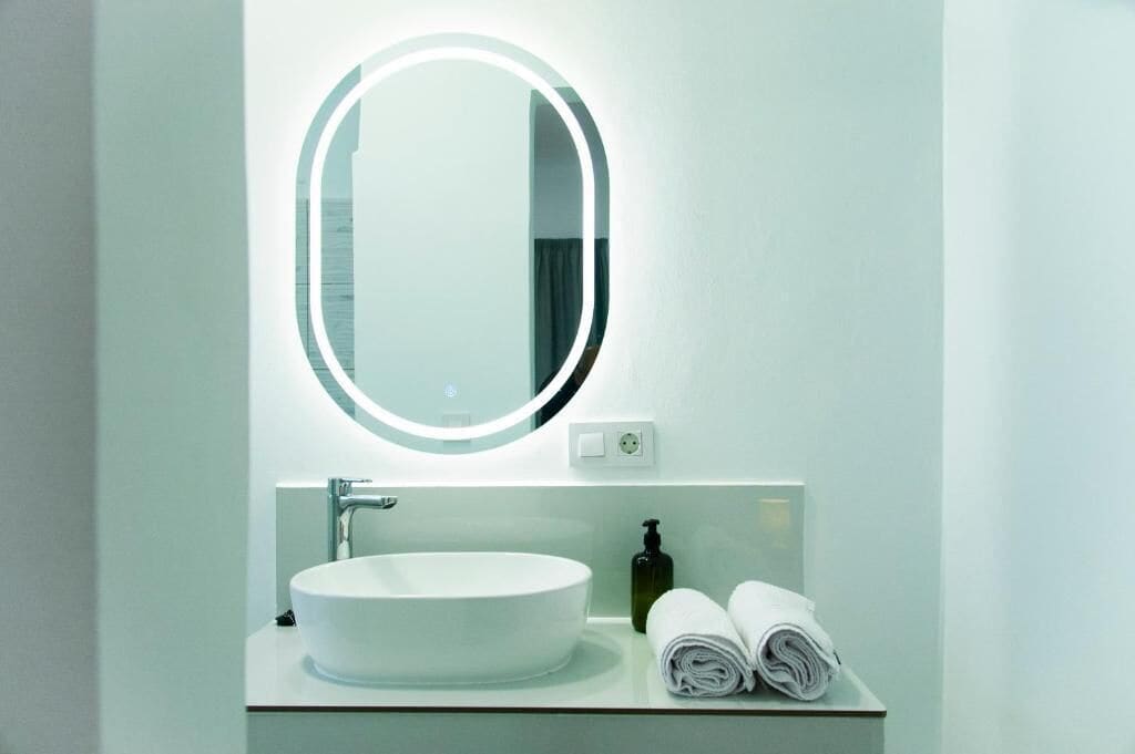 A bathroom with a round mirror, a white sink, a soap dispenser, and rolled towels on the counter.
