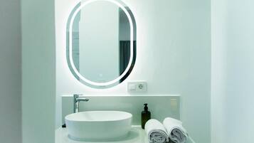 A bathroom with a round mirror, a white sink, a soap dispenser, and rolled towels on the counter.