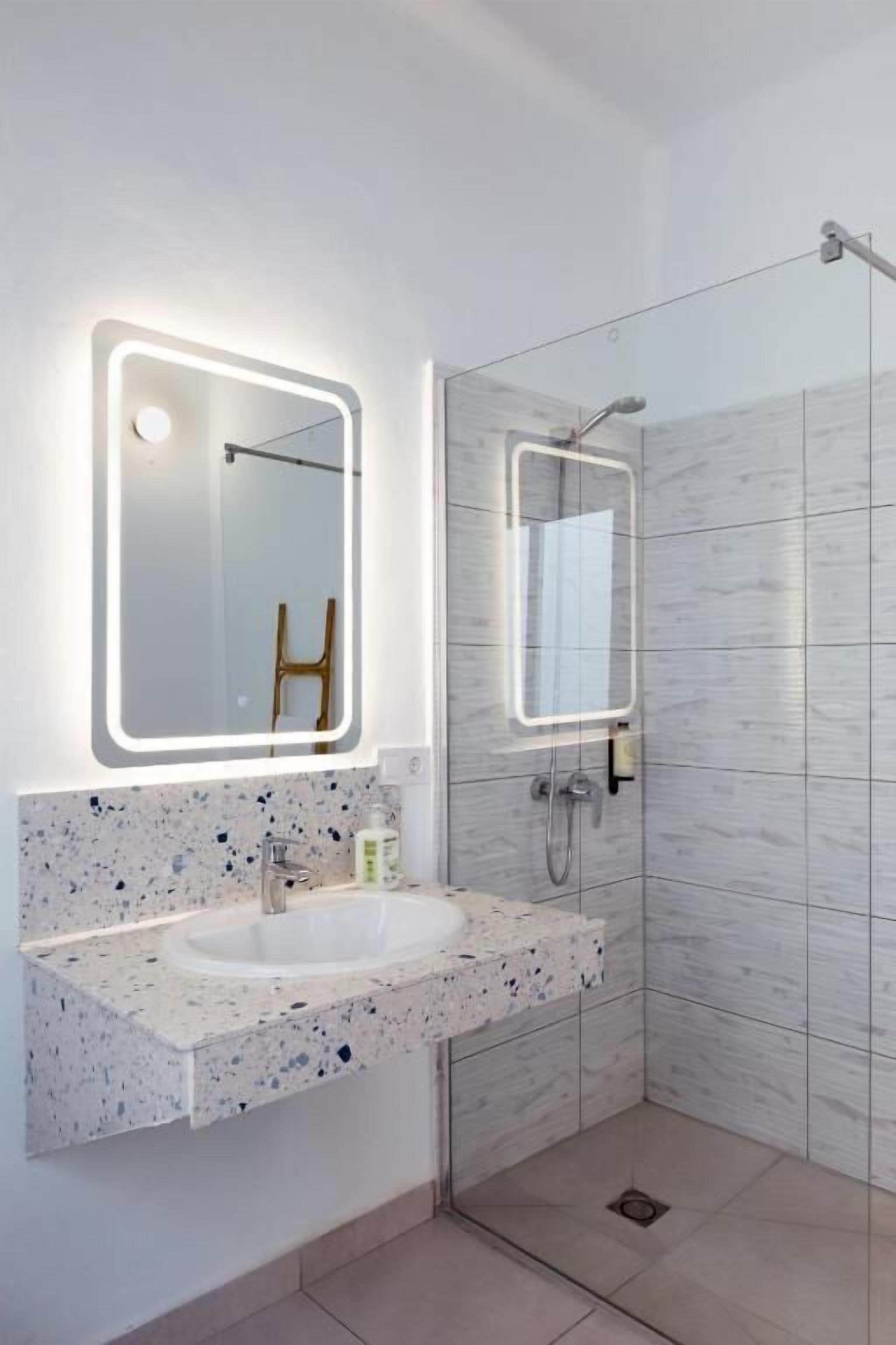 A modern bathroom with a granite countertop sink, a mirror with integrated lighting, and a shower area with a glass door.