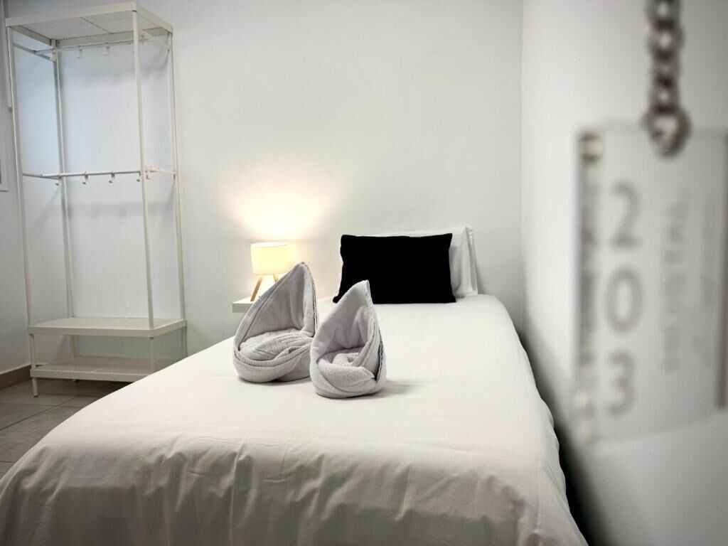 A hotel room with a bed, a white bedside table, a lamp, a black pillow, and a towel arrangement.