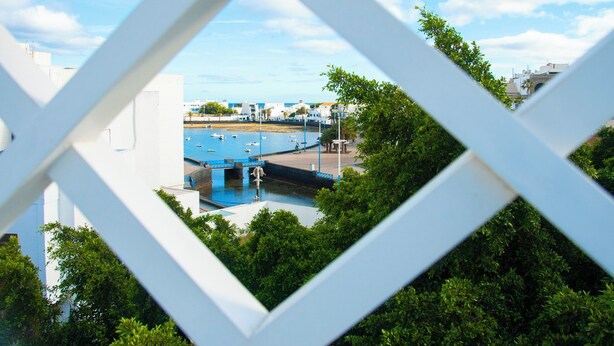 A view through a white geometric structure, revealing a marina with boats, a swimming pool, and buildings in the background. - Hostal España (Arrecife)