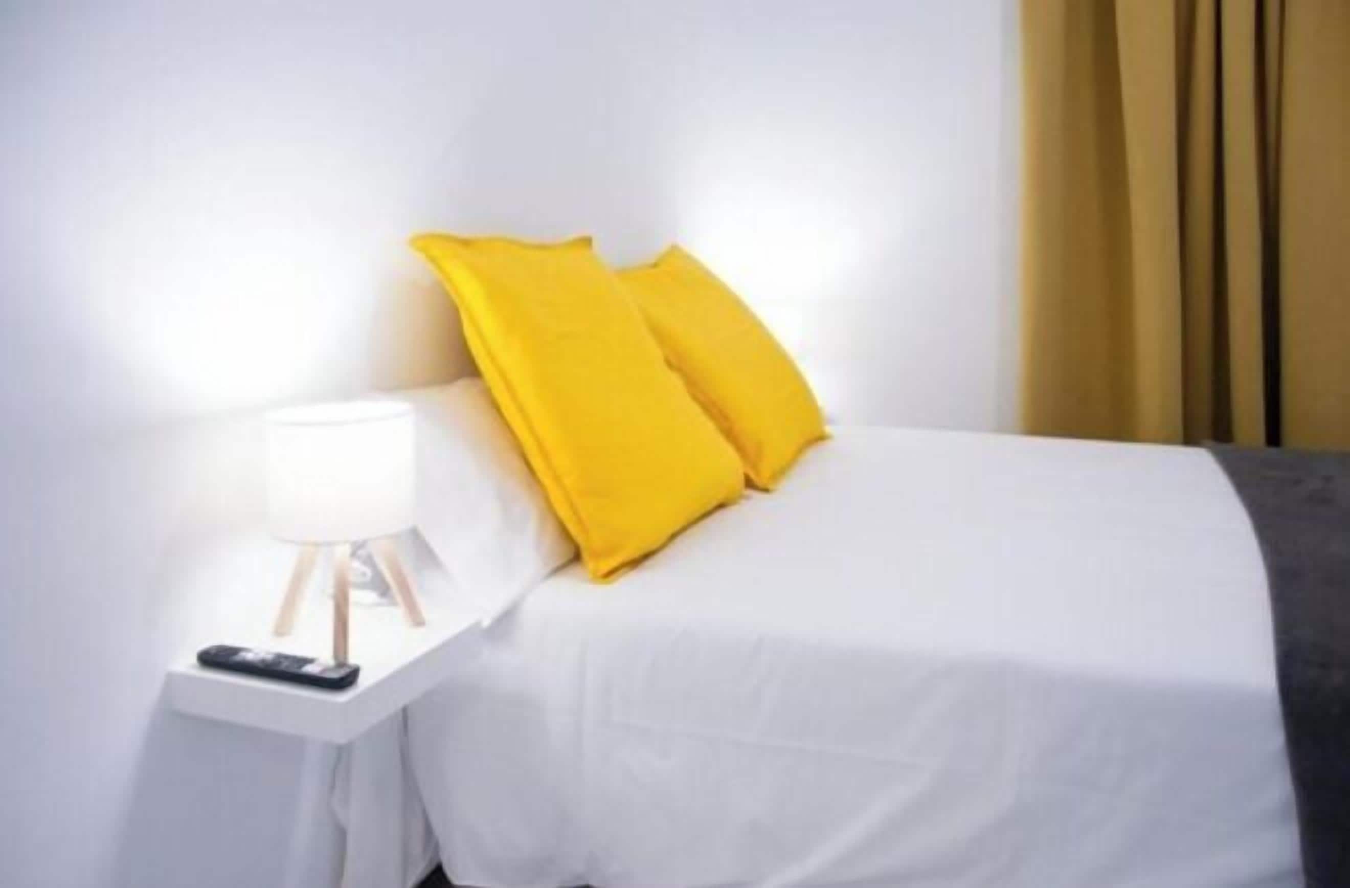 A bed with white bedding and two bright yellow pillows, a bedside table with a lamp, and a remote control.