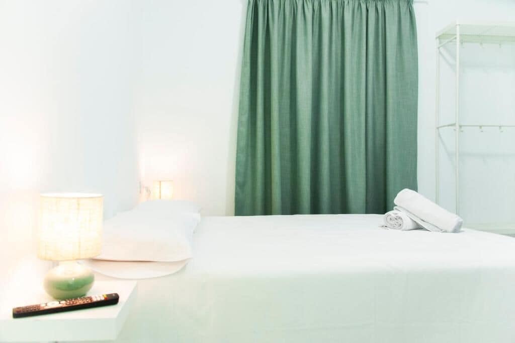 Una cama con ropa de cama blanca, una cortina verde, una lámpara de noche y una toalla enrollada sobre la cama.