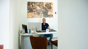 A person sitting at a desk with a computer, a phone, and a cup. There is a chair in front of the desk and a picture on the wall. - Hostal España (Arrecife)