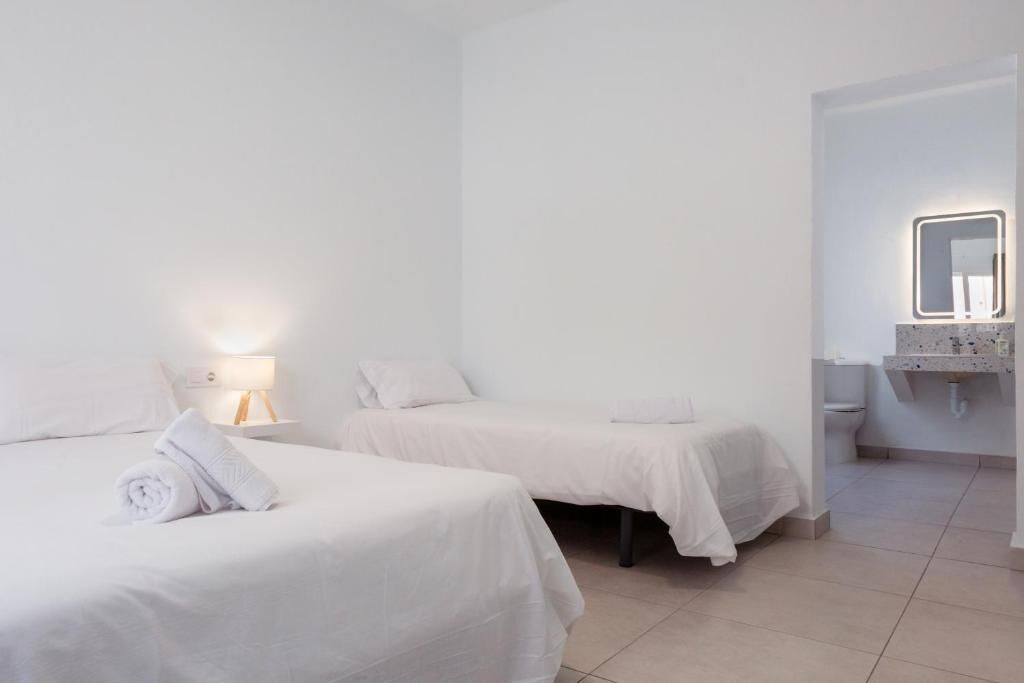 A hotel room with two beds, a bedside table with a lamp, a bathroom with a sink and mirror, and a toilet.
