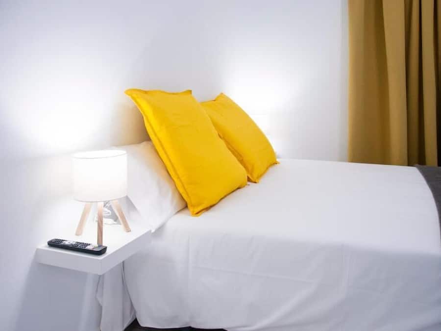 Una cama con ropa de cama blanca y dos almohadas amarillo brillante, una mesita de noche con una lámpara y un control remoto.
