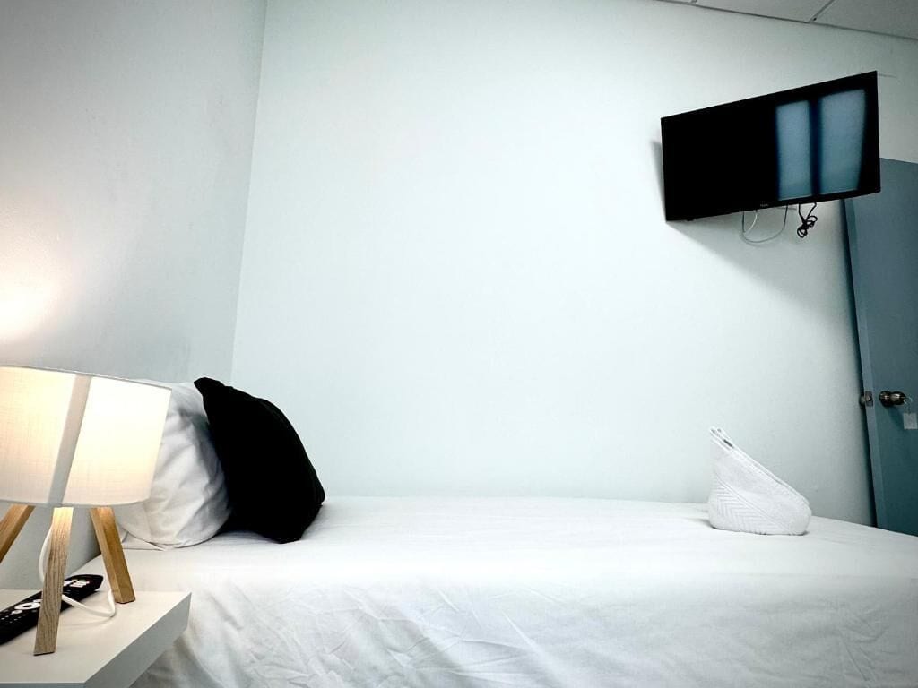 A hotel room with a bed, a flat-screen TV mounted on the wall, a bedside table with a lamp, and a white pillow.