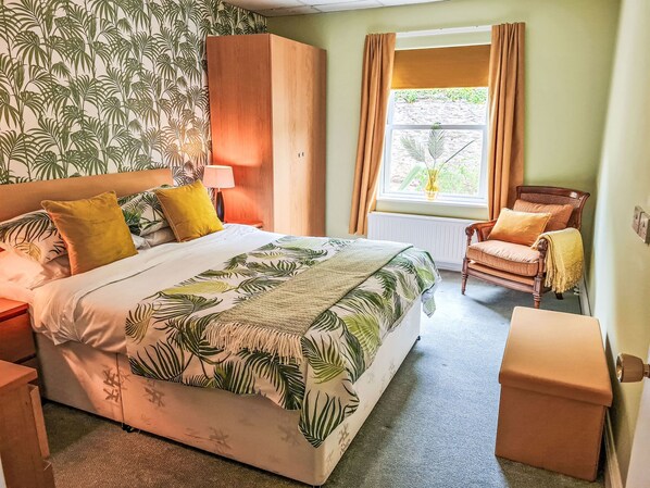 A room with a bed, a chair, a nightstand, a wardrobe, a window with curtains, and a wall with a leaf pattern. - The Mews by Interhome (Torquay)