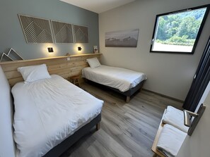Ein Zimmer mit zwei Betten, einem Holzkopfteil, einer Wandleuchte, einem gerahmten Bild und einem Fenster mit Blick auf Bäume.