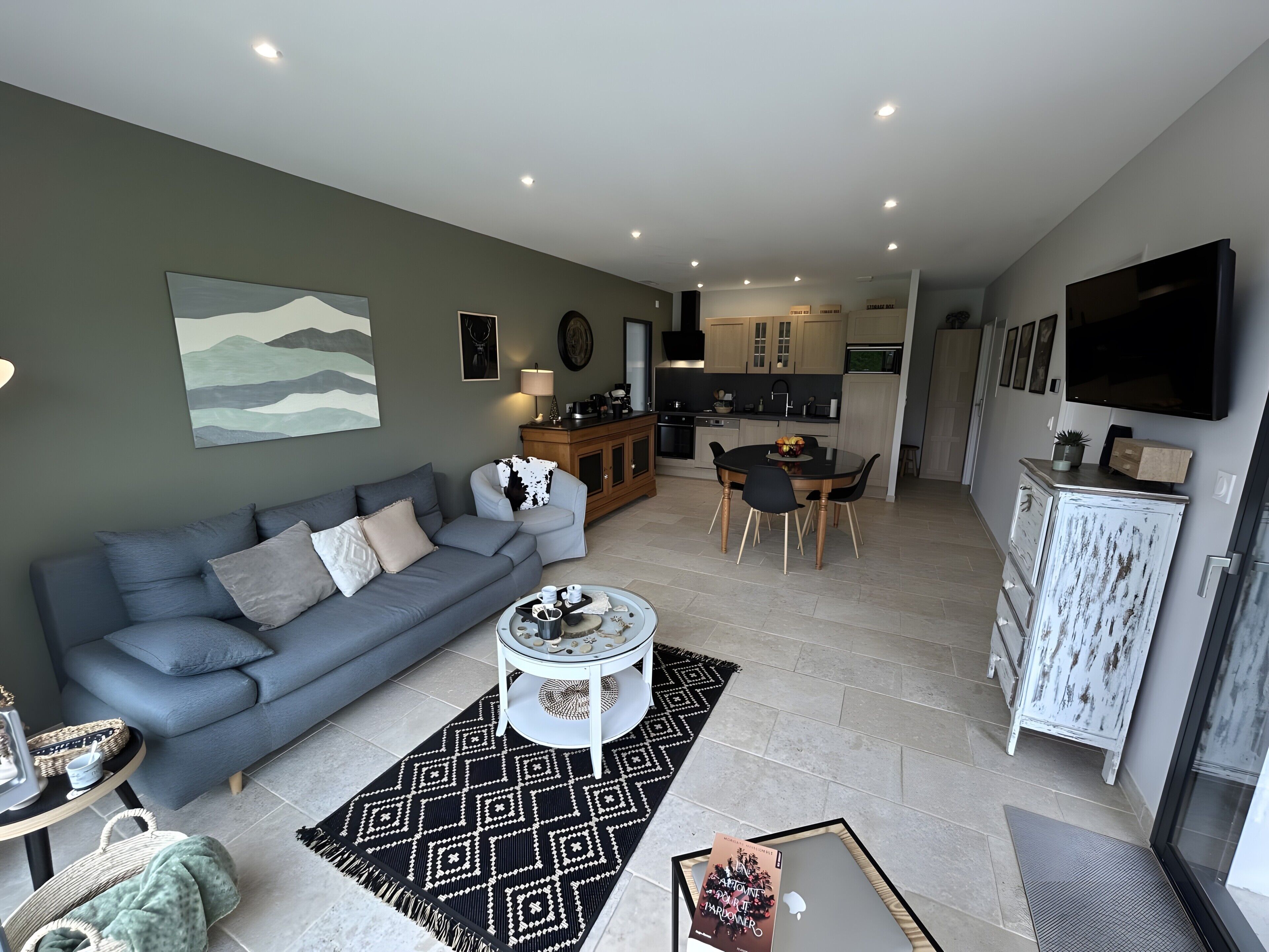 A modern living room with a grey sofa, a dining area with a black table, and a kitchen with wooden cabinets.