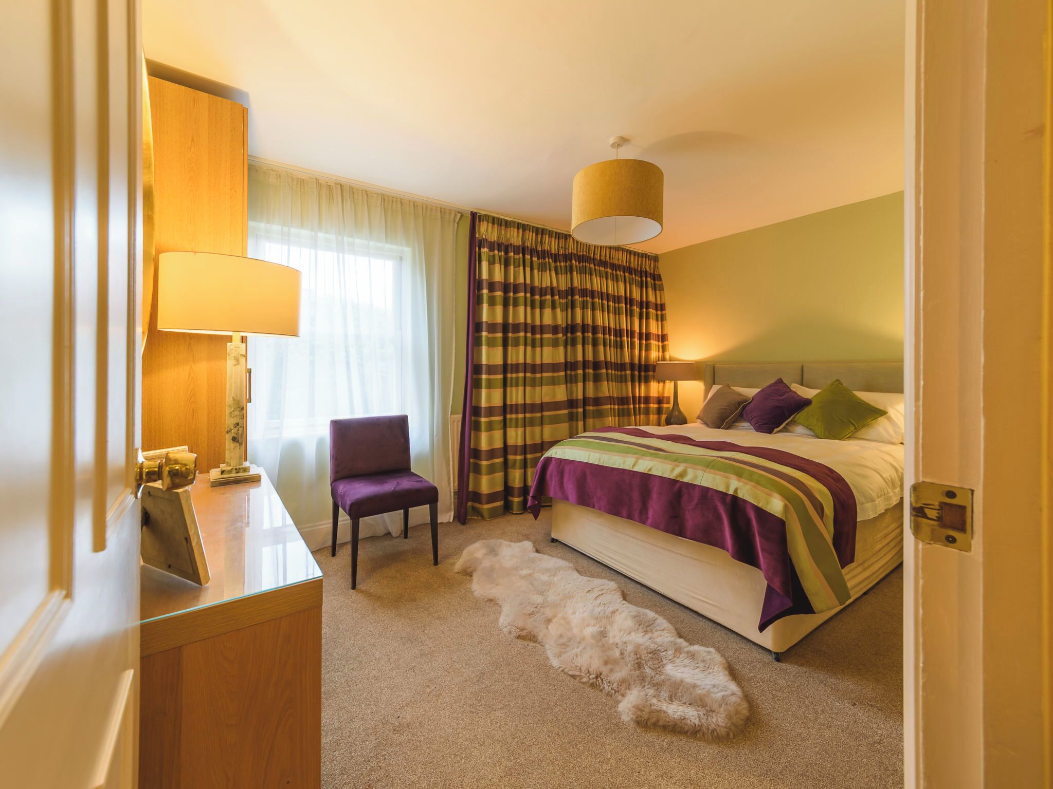 A hotel room with a bed, a chair, a lamp, a large window with curtains, and a carpet.