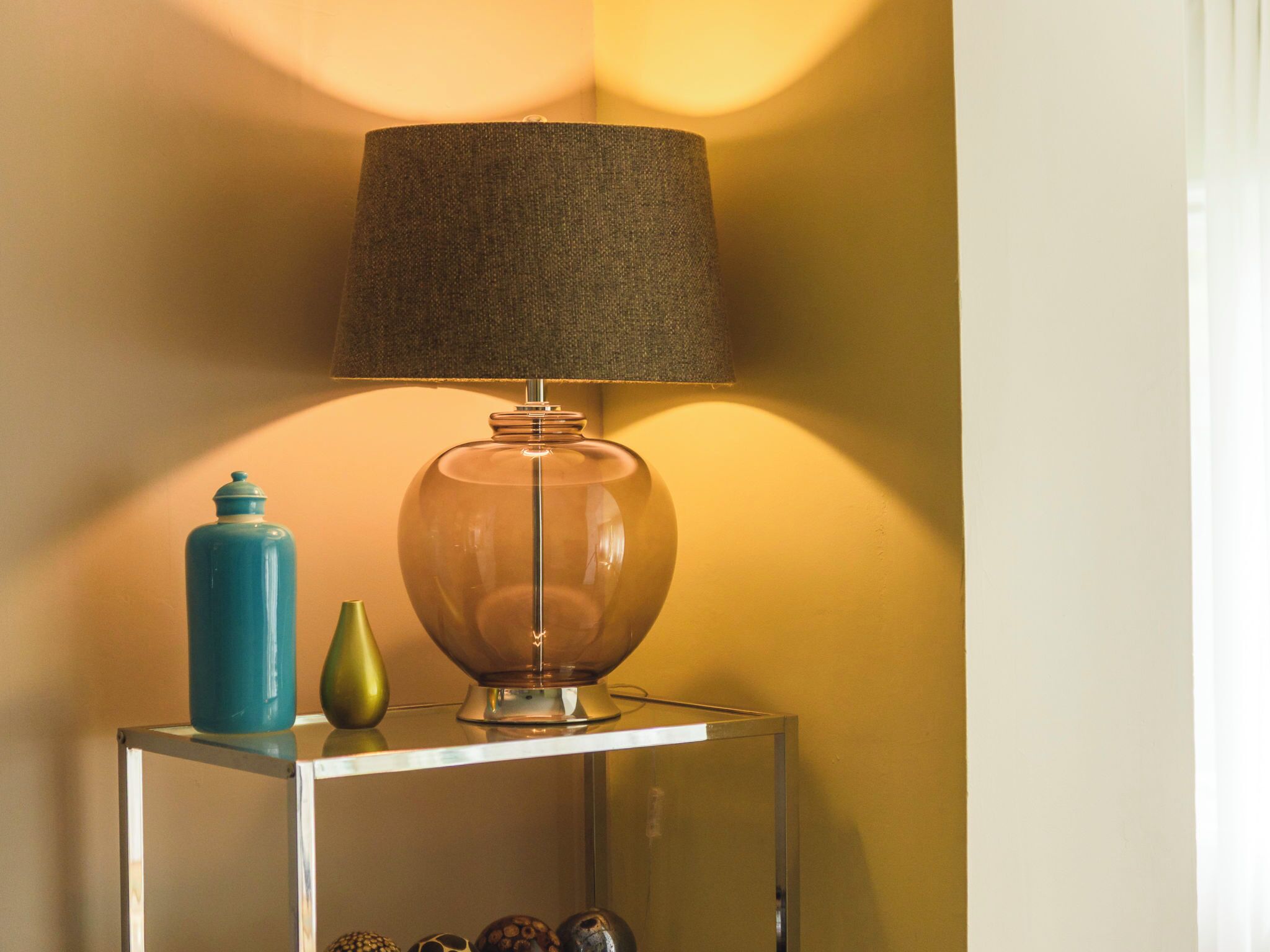Une lampe de table avec un abat-jour texturé, un vase turquoise et un vase vert, posés sur une table en verre transparent devant un mur jaune.