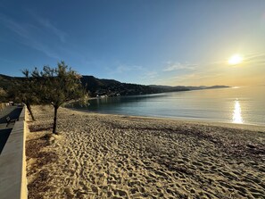 En strand med en asfalterad strandpromenad, träd och fri utsikt över havet.