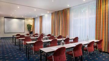 A conference room with tables, chairs, a projector screen, and a large window with curtains.