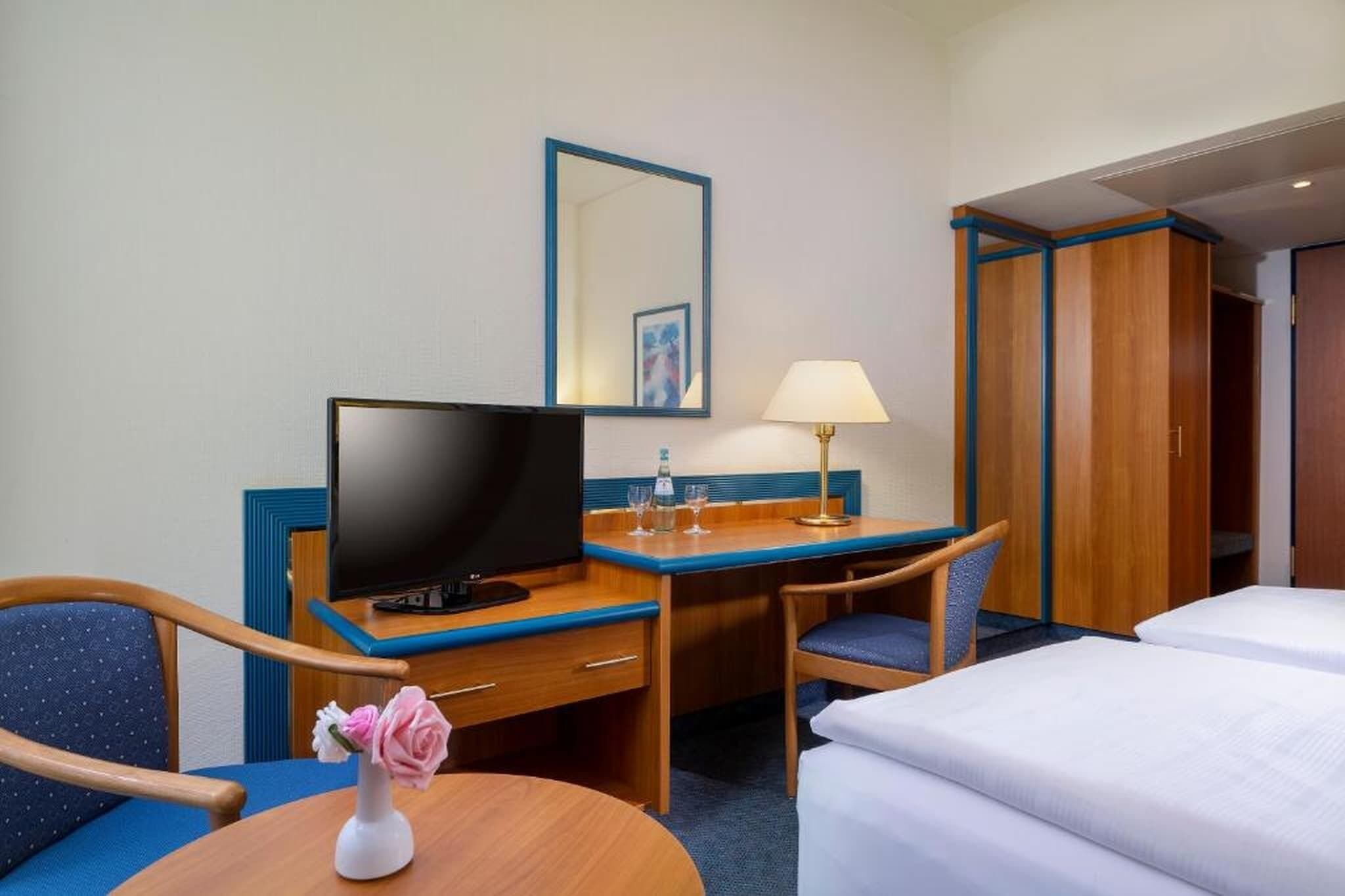 Ein Hotelzimmer mit Fernseher, Schreibtisch, zwei Stühlen, einem kleinen Tisch mit einer Vase und einem Bett.