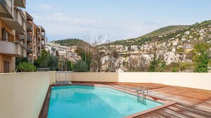 A swimming pool with a wooden deck, surrounded by buildings and a hilly landscape. - Apartment in Grasse Near Restaurants (Grasse)