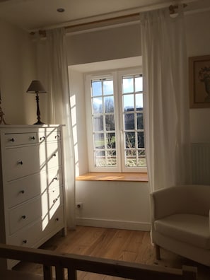 Ein Zimmer mit Fenster, Kommode, Stuhl und Lampe.