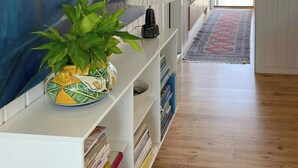 Un pasillo con una consola blanca, una planta en maceta y una lámpara. Se ven estanterías, una puerta y una vista a través de la puerta.