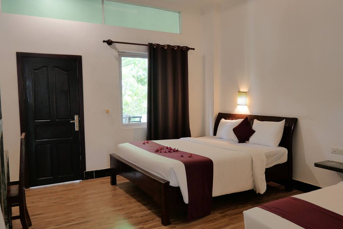 A hotel room with a bed, a bedside table, a window with curtains, a door, and a wooden floor.