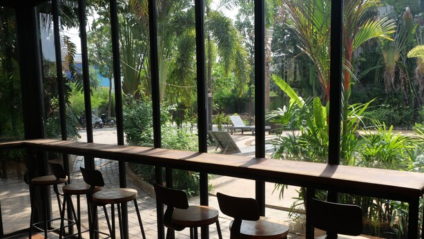 A bar area with stools and a view of a garden through large windows. - STC Hotel School (Siem Reap)