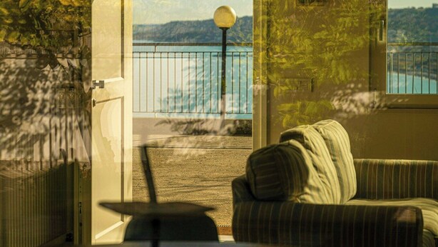 An open door leading to a balcony with a view of the sea, a round table with a chair, and a striped sofa.