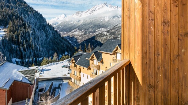 A wooden balcony with a view of snow-covered mountains and a village. - Alpine Comfort Apartment Near Ski-slopes (Modane)