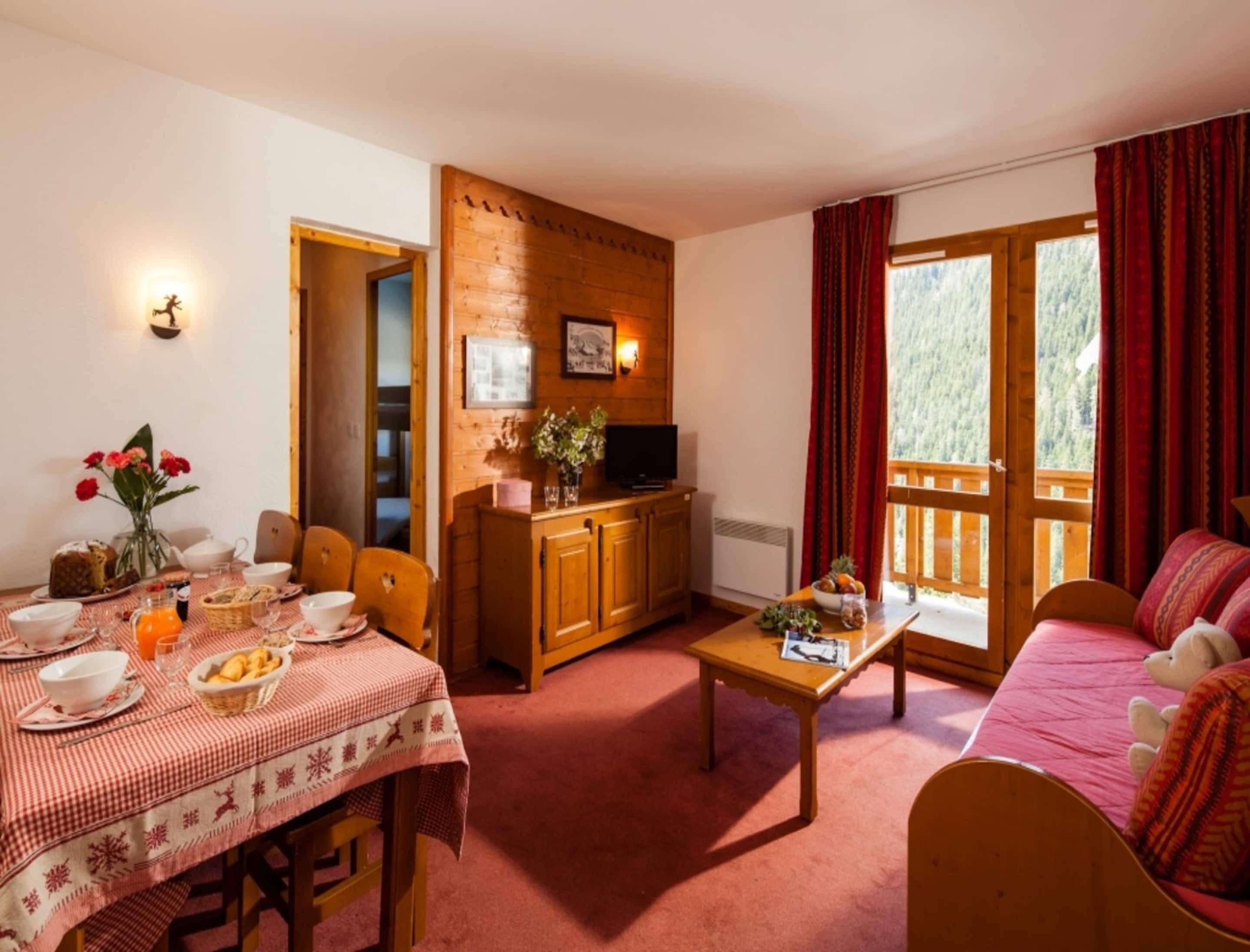 A cozy room with a dining table set for breakfast, a sofa with a stuffed toy, a wooden cabinet, and a large window with curtains.