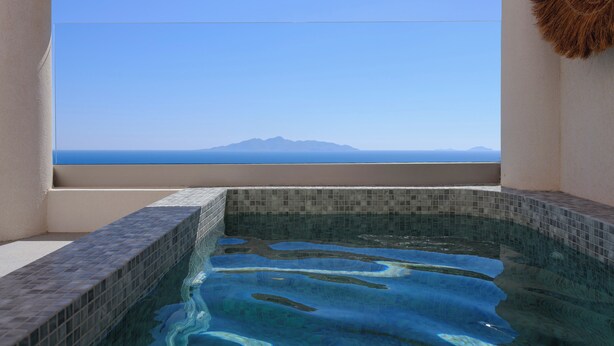 On aperçoit clairement une piscine dont le bord est recouvert de carreaux, et une fenêtre donne sur une montagne et la mer, situées au loin.