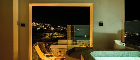 A modern hotel room with a glass-enclosed balcony, a hot tub, and a view of a cityscape at night.