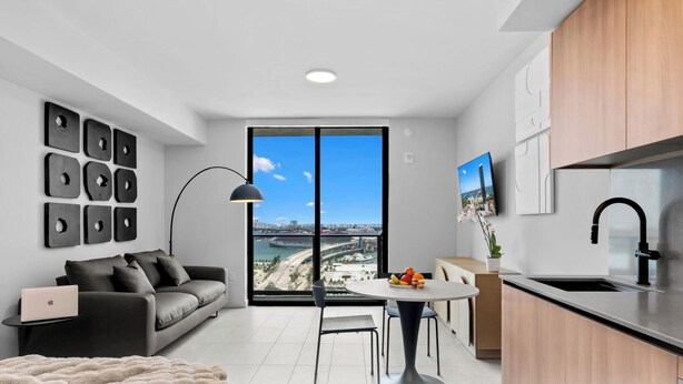 A modern apartment with a sofa, dining table, and kitchen area, featuring a large window with a city view. - 501 First Studio with Luxury Amenities (Miami)