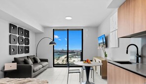 A modern apartment with a sofa, dining table, and kitchen area, featuring a large window with a city view. - 501 First Studio with Luxury Amenities (Miami)