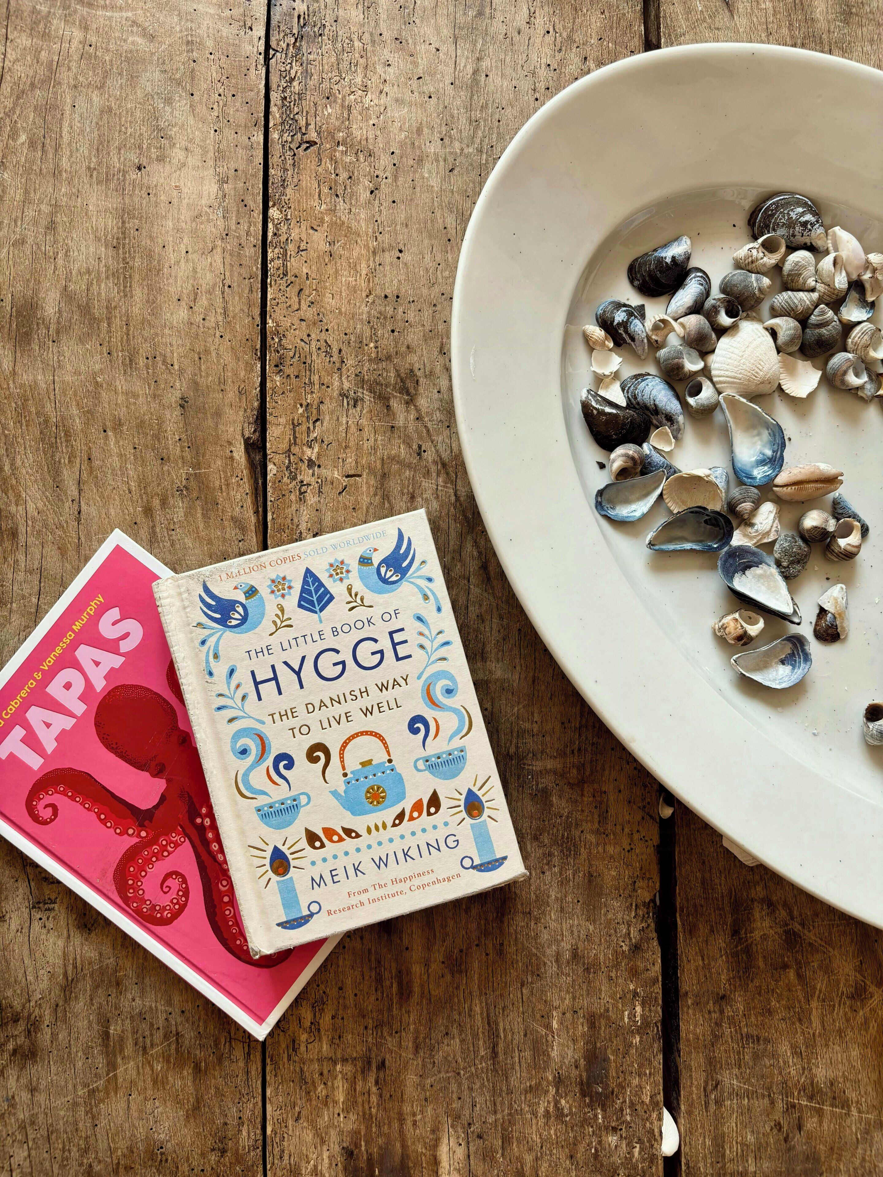 A wooden table with a white bowl containing seashells, a book titled 'The Little Book of Hygge,' and another book with a red cover.