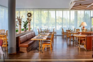 Un'ampia sala da pranzo con diversi tavoli e sedie, un divano centrale e grandi finestre con tende leggere.