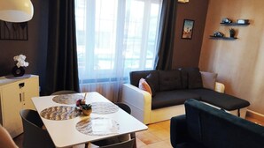 Huoneessa on ruokapöytä, tuoleja, sohva, musta nojatuoli, lamppu, ikkuna, jossa on verhot, sekä seinä, jossa on taulu ja hyllyjä.
